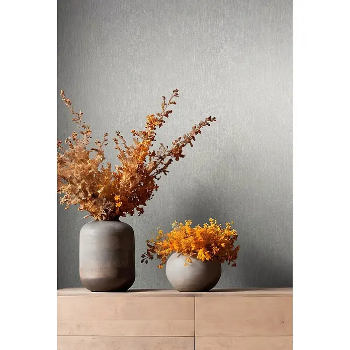 Marburg Ritus Vliestapete Uni I - Grau, Uni, 10,05 x 0,53 m - Zwei Keramikvasen mit Trockenblumen auf einem hellen Holzschrank. Die hohe Vase enthält goldbraunes Laub, die runde Vase leuchtend orangefarbene Blüten.