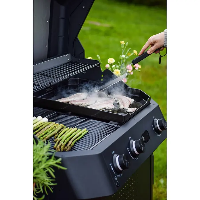Cozze Grillgutwender - Länge: 42 cm, Edelstahl/Kunststoff - Schwarzer Gasgrill kocht Fisch auf einer Platte und Spargel auf Rosten, eine Hand benutzt einen Spatel.