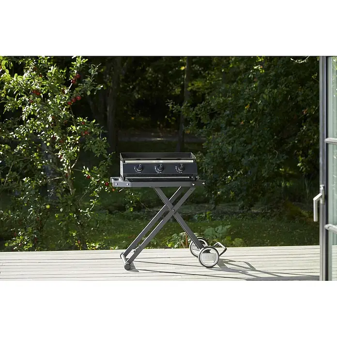Cozze Klapptisch - B x T x H: 104,5 x 62,5 x 78 cm, Stahl - Eine schwarze Cozze Grillplatte auf einem grauen, zusammenklappbaren Ständer mit Rädern steht auf einer Holzterrasse im Freien, umgeben von Bäumen mit roten Äpfeln.