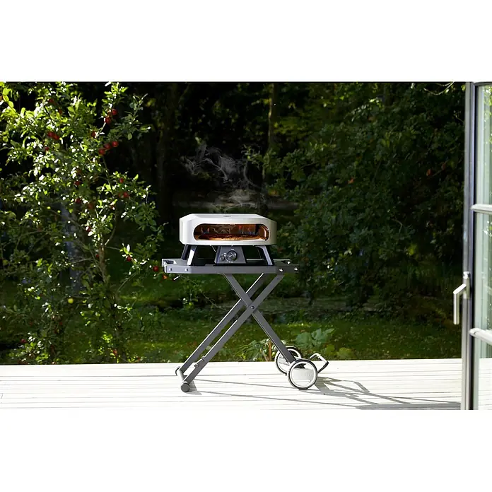 Cozze Klapptisch - B x T x H: 104,5 x 62,5 x 78 cm, Stahl - Weißer Outdoor-Pizzaofen auf grauem Rollständer, backt eine Pizza mit sichtbarem Rauch im sonnigen Garten.