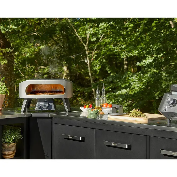 Cozze Pizzaofen Classic 13 - Grillfläche (B x T): 34 x 34 cm, 6 kW - Moderne schwarze Außenküche mit grauem Pizzaofen, der eine Pizza backt. Zubereitungsbereich mit Gemüse und Ölen sichtbar.