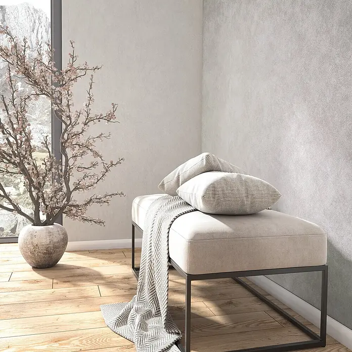 Marburg Whisper Vliestapete Putzoptik - Grau-Weiß, Sonstiges, 10,05 x 0,53 m - Beige gepolsterte Bank mit dunklem Metallgestell, akzentuiert mit gestreiften Kissen und einer grauen Decke in einem hellen Raum.