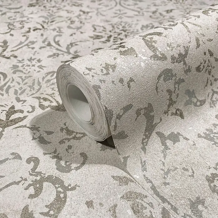 Marburg City Glow Vliestapete Ornamental - Beige, Ornament, 10,05 x 0,53 m - Teilweise abgerollte Tapetenrolle mit metallischem Damastmuster in Silber auf hellem Hintergrund.