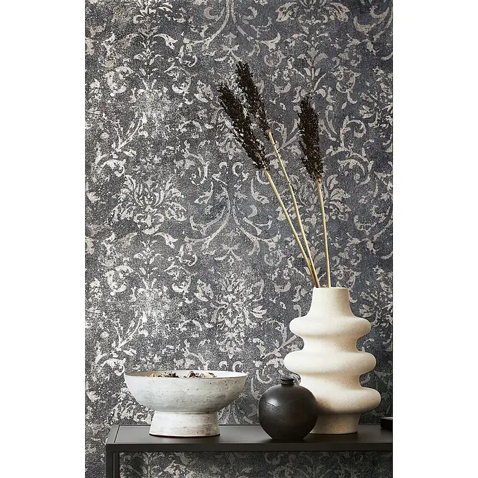Marburg City Glow Vliestapete Ornamental - Schwarz-Silber, Ornament, 10,05 x 0,53 m - Regal-Dekor: strukturierte cremefarbene Vase mit Trockenblumen, weiße Schale, schwarze Kugel vor gemusterter Tapete.