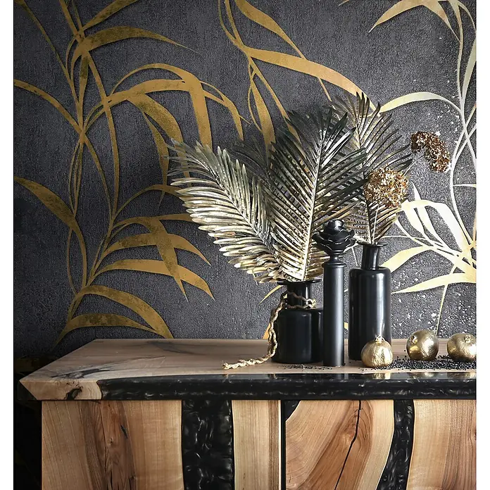 Marburg City Glow Vliestapete Uni II - Schwarz-Gold, Uni, 10,05 x 0,53 m - Kommode aus Holz mit dunklen Harzakzenten, dekoriert mit schwarzen Vasen und goldenen Palmblättern. Vor einer dunklen, goldgemusterten Wand.