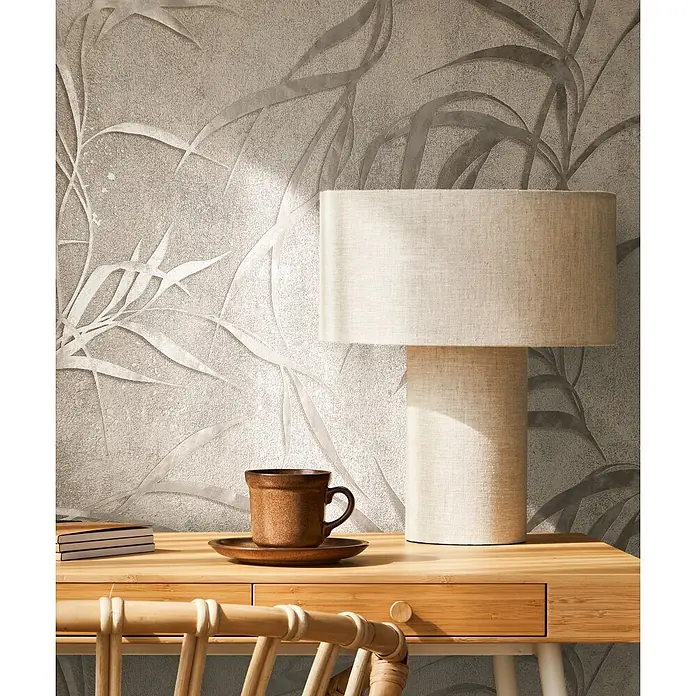 Marburg City Glow Vliestapete Uni II - Grau, Uni, 10,05 x 0,53 m - Eine beige Leinenlampe, eine braune Keramiktasse und Bücher auf einem hellen Holzschreibtisch vor einer gemusterten Wand.