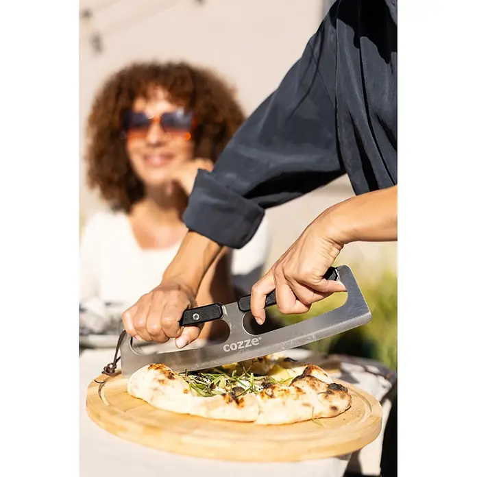 Cozze Pizzaschneider - Stahl, Länge: 32 cm - Eine Person benutzt einen silbernen Pizzaschneider mit schwarzen Griffen, um eine Pizza auf einem Holzbrett zu schneiden. 'cozze' ist auf der Klinge sichtbar.