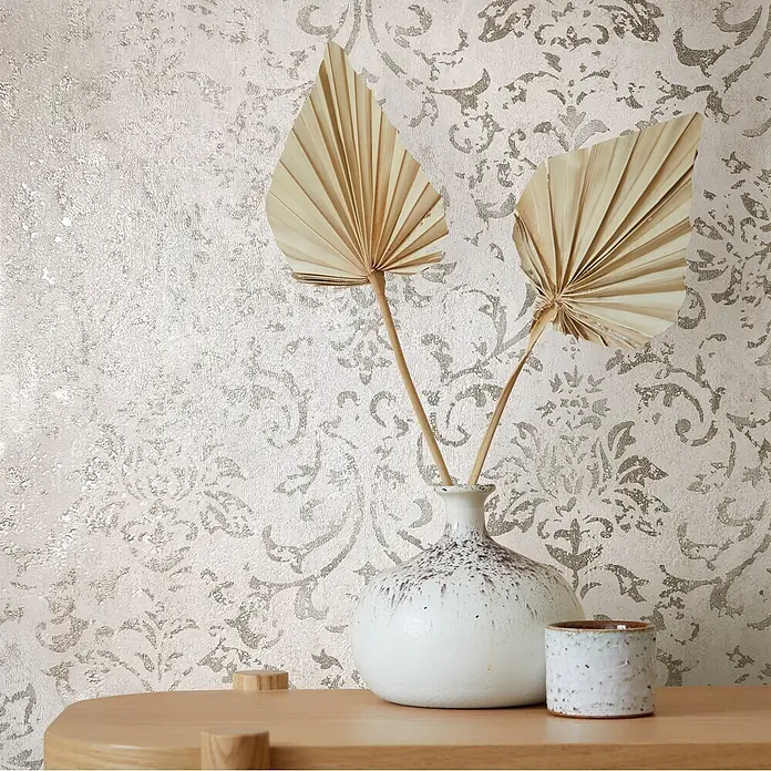 Marburg City Glow Vliestapete Ornamental - Beige, Ornament, 10,05 x 0,53 m - Strukturierte hellbeige Tapete mit metallischem Damastmuster, in einer Raumeinstellung mit Vase und getrockneten Palmblättern auf einem Holztisch.