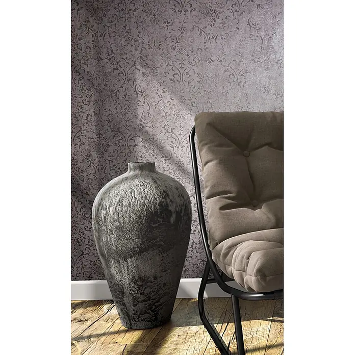 Marburg City Glow Vliestapete Ornamental - Braun, Ornament, 10,05 x 0,53 m - Eine große, graue, strukturierte Vase steht auf einem Holzfußboden neben einem beigen Polsterstuhl mit schwarzem Gestell.