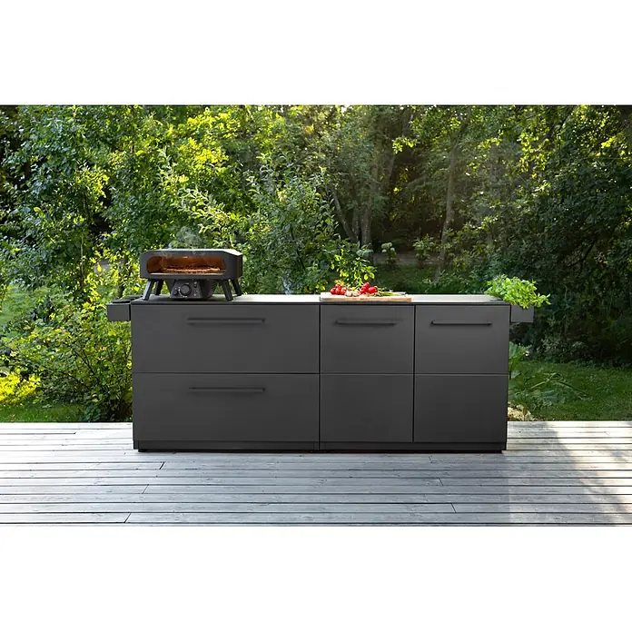 Cozze Pizzaofen Black Edition 17" mit drehbarem Pizzastein - Durchmesser Grillfläche: 43 cm, 8 kW - Dunkelgraue Outdoor-Küchenzeile mit Pizzaofen und Vorbereitungsbereich auf einer Holzterrasse.