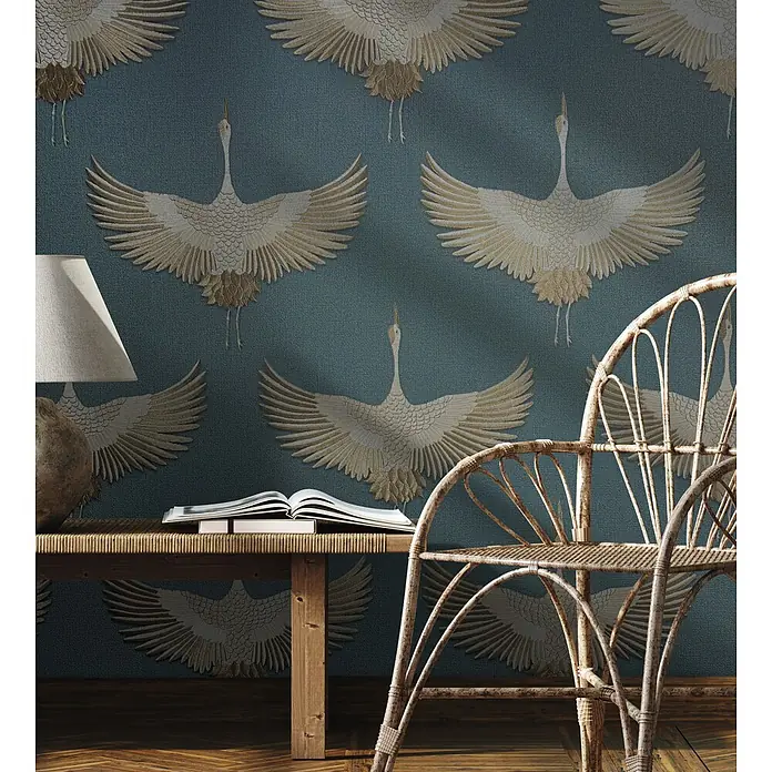 Marburg Kumano Vliestapete Grafik - Blau, Grafisch, 10,05 x 0,53 m - Türkisfarbene Tapete mit eleganten goldenen Kranichen schmückt eine Wohnzimmerwand hinter einem Holztisch und einem Rattansessel.