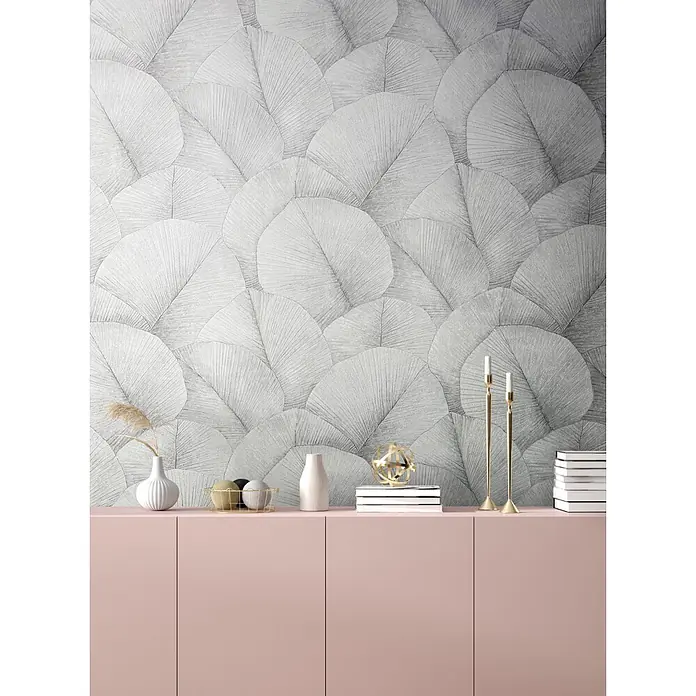 Marburg Kumano Vliestapete Floral - Hellgrau, Floral, 10,05 x 0,53 m - Rosa Unterschrank mit Dekoartikeln wie Vasen, Büchern und goldenen Kerzenhaltern, vor einer grauen Wand mit Blattmuster.