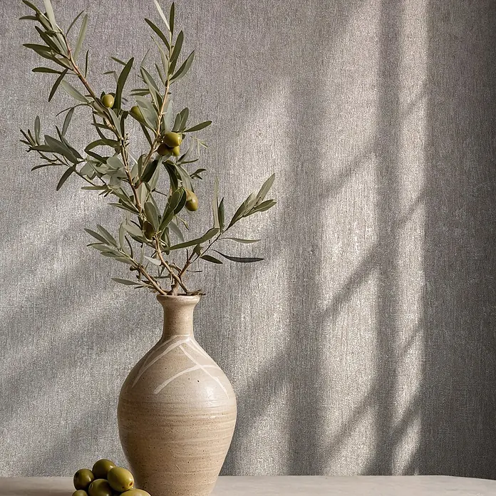 Marburg Ritus Vliestapete Uni - Grau, Uni, 10,05 x 0,53 m - Olivenzweige mit grünen Oliven in einer beigen Keramikvase auf einem Tisch vor strukturierter Wand mit Lichtmustern.