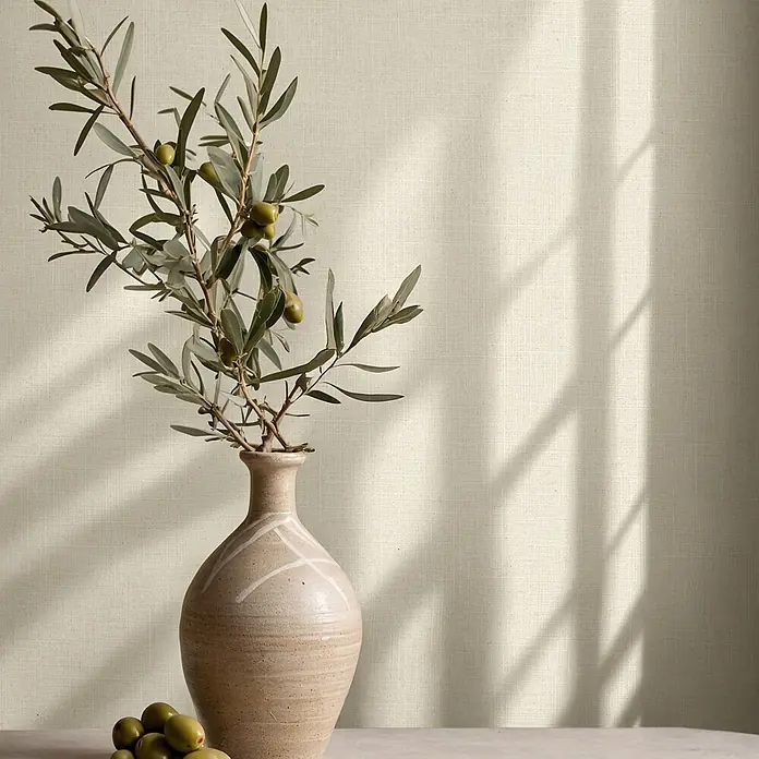 Marburg Boho Vliestapete Uni - Beige, Uni, 10,05 x 0,53 m - Olivenzweig mit grünen Oliven in beiger Keramikvase auf Tisch vor strukturierter Wand mit Licht und Schatten.