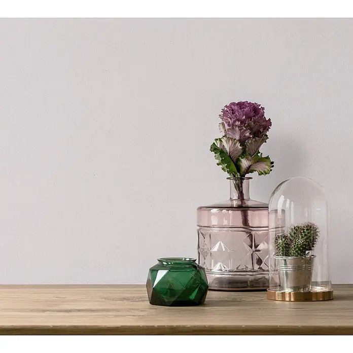 Marburg Vliestapete Novamur Oriana Uni - Weiß-Rosa, Uni, 10,05 x 0,53 m - Eine Sammlung dekorativer Glasartikel: eine rosa Vase mit Pflanze, eine grüne facettierte Schale und ein Kaktus unter einer Glaskuppel auf einem Holztisch.