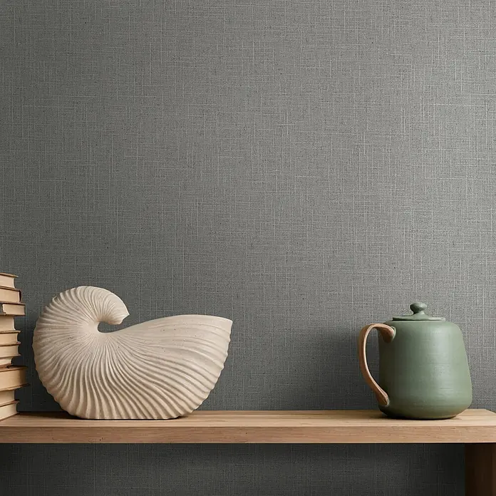 Marburg Boho Vliestapete Uni II - Grau-Braun, Uni, 10,05 x 0,53 m - Ein Holzregal präsentiert Bücher, eine cremefarbene Nautilus-Muschel und eine grüne Keramikkanne vor einer strukturierten Wand.