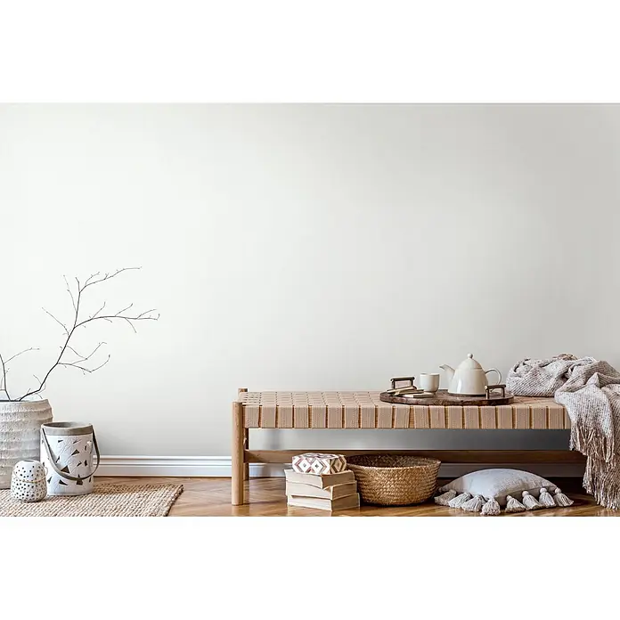 Marburg Vliestapete Novamur Oriana Uni - Creme, Uni, 10,05 x 0,53 m - Lange Holzbank mit geflochtener Sitzfläche, dekoriert mit Teeservice und Decke in einem hellen, minimalistischen Interieur.