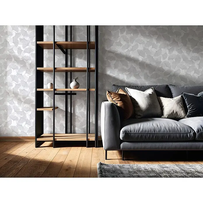 Marburg Boho Vliestapete Floral - Grau, Floral, 10,05 x 0,53 m - Ein modernes Wohnzimmer zeigt ein schwarz-holzfarbenes Bücherregal vor einer gemusterten Wand, neben einem grauen Sofa mit Zierkissen.