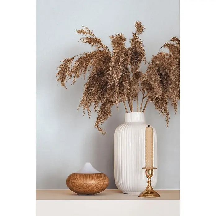 Marburg Vliestapete Novamur Oriana Uni - Blau, Uni, 10,05 x 0,53 m - Ein Diffusor aus Holz, eine weiße geriffelte Vase mit getrocknetem Pampasgras und eine Bienenwachskerze in einem Messinghalter auf einem hellen Regal.