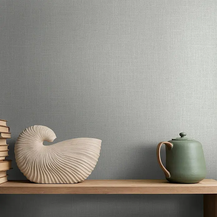 Marburg Boho Vliestapete Uni I - Beige-Grau, Uni, 10,05 x 0,53 m - Dezente, gewebte hellgraue Wandverkleidung, dekoriert mit einem Naturholzregal mit Büchern, einer Nautilusschale und einer grünen Teekanne.