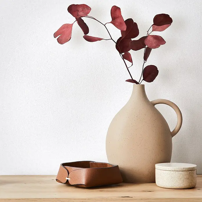 Marburg Ritus Vliestapete Putzoptik I - Weiß, Sonstiges, 10,05 x 0,53 m - Eine beige Vase mit roten Blättern, ein braunes Lederkörbchen und eine gesprenkelte Keramikdose auf einem Holztisch.