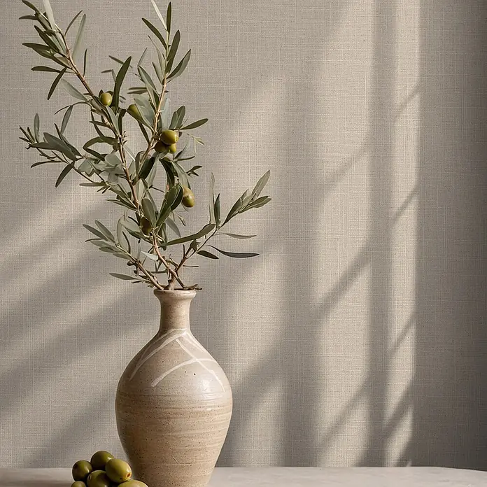 Marburg Boho Vliestapete Uni II - Beige, Uni, 10,05 x 0,53 m - Olivenzweige mit grünen Oliven in einer hellbeigen Keramikvase, mit losen Oliven auf einem Tisch vor einer strukturierten Wand mit Sonnenschatten.