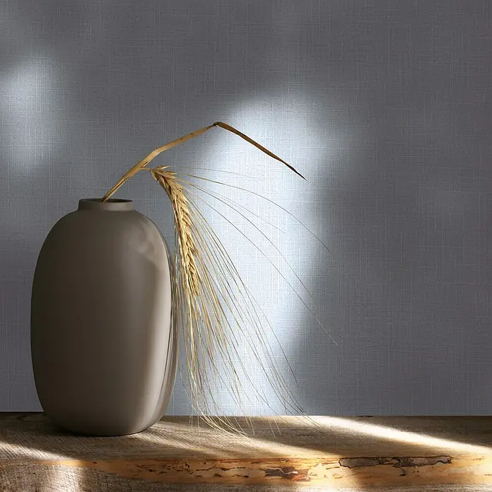 Marburg Boho Vliestapete Uni I - Blau-Grau, Uni, 10,05 x 0,53 m - Eine matte graue ovale Vase mit goldenen Weizenähren auf einem Holzuntergrund, gebadet in sanftes Licht.