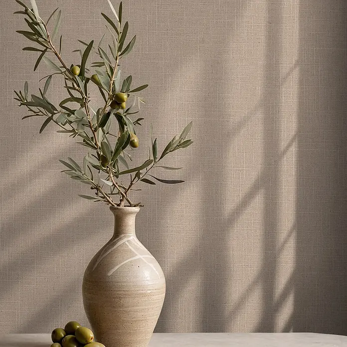 Marburg Boho Vliestapete Uni II - Taupe, Uni, 10,05 x 0,53 m - Olivenzweig mit grünen Oliven in heller Keramikvase auf Tisch vor texturiertem beigem Hintergrund mit Schatten.