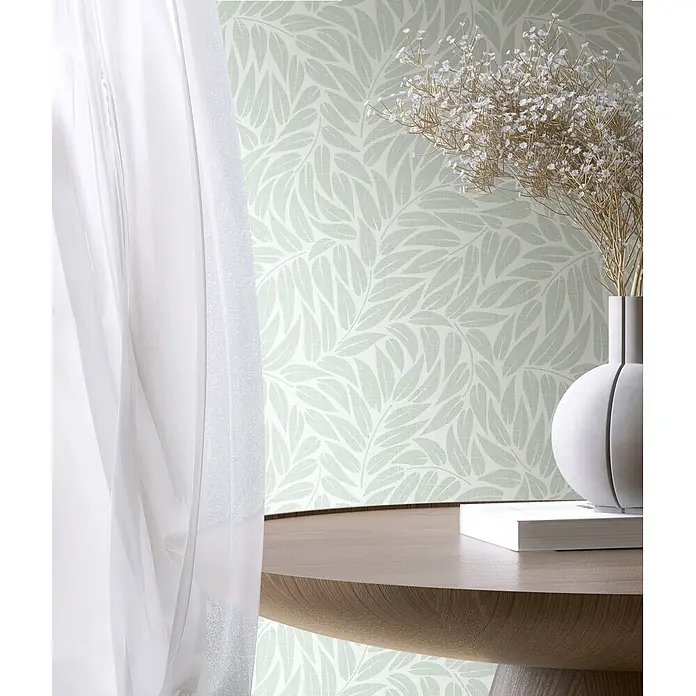Marburg Boho Vliestapete Floral I - Grün, Floral, 10,05 x 0,53 m - Hellgrüne Tapete mit dezentem Blattmuster und Leinenstruktur, an einer Wand hinter einem Holztisch mit Trockenblumen und einem Buch.
