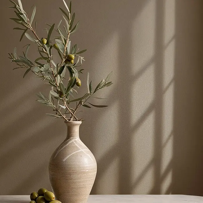 Marburg Vliestapete Novamur Naomi Uni I - Braun, Uni, 10,05 x 0,53 m - Strukturierte beige Vase mit Olivenzweig und grünen Oliven, einige auf dem Tisch, vor strukturierter brauner Wand mit Schatten.
