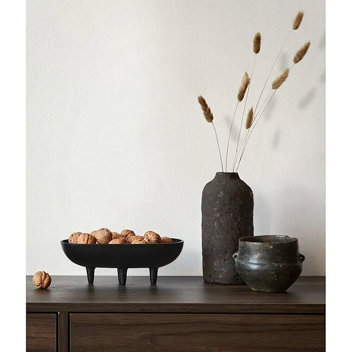 Marburg Whisper Vliestapete Uni - Weiß-Beige, Uni, 10,05 x 0,53 m - Eine dunkle Holzkommode präsentiert eine schwarze Schale mit Walnüssen, eine hohe schwarze strukturierte Vase mit Trockengras und einen kleinen rustikalen Topf.