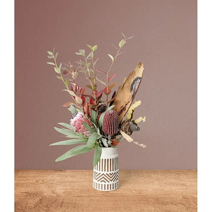 Marburg Vliestapete Novamur Naomi Uni I - Rot-Braun, Uni, 10,05 x 0,53 m - Eine weiße Vase mit braunen geometrischen Mustern enthält ein Trockenblumenarrangement mit Protea, Banksia, Eukalyptus und Holz auf einem Holztisch.