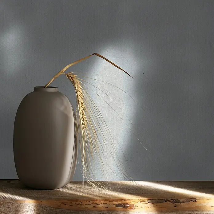 Marburg Whisper Vliestapete Uni I - Grau, Uni, 10,05 x 0,53 m - Eine längliche, mattgraue Vase mit einer getrockneten Weizenähre, auf einer rustikalen Holzfläche mit natürlichem Licht.