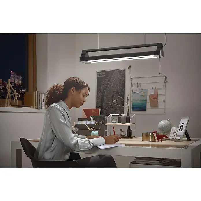 Osram LED-Deckenleuchte Office Line Double Portable - L x B x H: 60 x 11,9 x 10,22 cm, Schwarz, Kaltweiß - Eine schwarze, rechteckige Pendelleuchte hängt über einem Schreibtisch und beleuchtet eine schreibende Frau. Sie bietet fokussiertes Arbeitslicht.