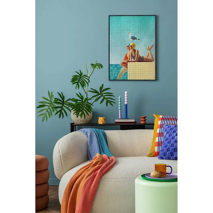 Accent Alurahmen Puzzle Frame - 50 x 70 cm, Schwarz - Ein gemütliches Wohnzimmer mit cremefarbenem Bouclé-Sofa, bunten Decken und Kissen, neben einer großen Topfpflanze und einem gerahmten Puzzlebild.