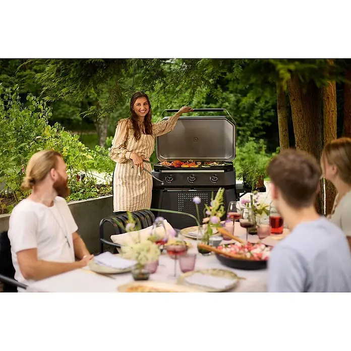 Cozze Plancha-Grill 800 - Grillfläche (B x T): 80 x 50 cm, 7,5 kW, Schwarz - Schwarz-silberner Grill, von einer Frau zum Garen von Speisen im Freien genutzt, mit Gästen am Tisch.