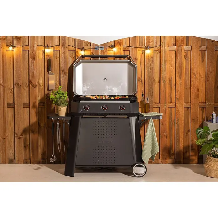 Cozze Rollwagen - B x T x H: 146 x 70 x 74,5 cm, Stahl, Schwarz, Passend für: Cozze Plancha Grill 800 - Schwarzer Cozze Gas-Plancha-Grill mit offener Edelstahlhaube, auf dem Speisen zubereitet werden, mit Seitenablagen und Rollen, vor einem Holzzaun.