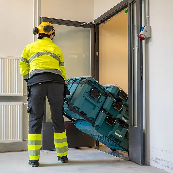 Makita Werkzeugkasten Maktrak Trolley L - Kunststoff, L x B x H: 101,1 x 56,5 x 29,5 cm - Arbeiter transportiert stapelbare türkisfarbene Makita-Werkzeugkisten auf einem Rollwagen durch eine Tür.