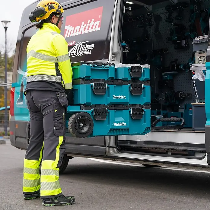 Makita Werkzeugkasten Maktrak Trolley L - Kunststoff, L x B x H: 101,1 x 56,5 x 29,5 cm - Türkis-schwarze Makita-Werkzeugboxen, gestapelt und rollbar, werden von einem Arbeiter in einen Transporter geladen.