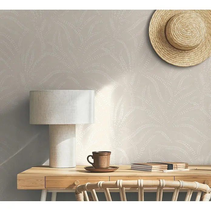 Marburg Vliestapete Novamur Oriana Floral - Beige, Floral, 10,05 x 0,53 m - Hellbeigefarbene Tapete mit weißem Farnmuster, die eine dezente, natürliche Kulisse für einen Schreibtisch mit Lampe, Tasse und Strohhut bildet.