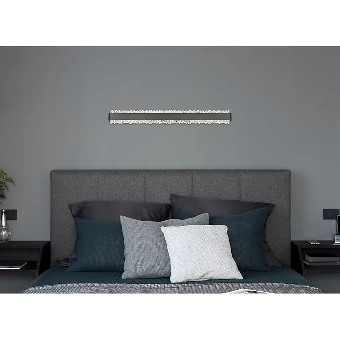 Globo LED-Wandleuchte Stalagmita - 18 W, Neutralweiß - Lineare Wandleuchte mit dunklem Metallsockel und funkelnden Kristallelementen, horizontal über einem Bett montiert.