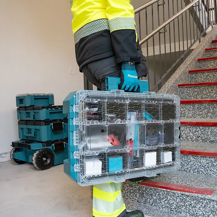 Makita Organizer Maktrak M - L x B x H: 56,2 x 38,7 x 16,2 cm, Anzahl Fächer: 11 Stk. - Person trägt durchsichtigen, türkisfarbenen Werkzeugorganizer mit Kleinteilen, gestapelte Boxen im Hintergrund.