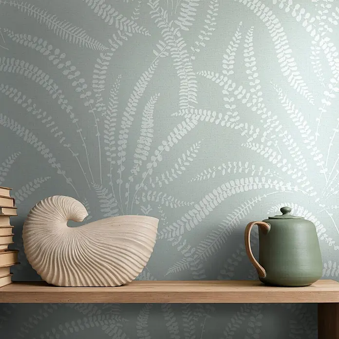 Marburg Vliestapete Novamur Oriana Floral - Grün, Floral, 10,05 x 0,53 m - Helle grüne Tapete mit weißem Farnmuster. Ein Holzregal trägt Bücher, eine Nautilus-Skulptur und eine grüne Teekanne.