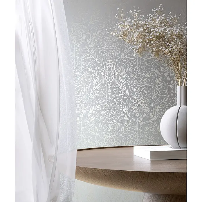Marburg Vliestapete Novamur Oriana Floral I - Grau-Blau, Floral, 10,05 x 0,53 m - Transparente weiße Vorhänge neben hellgrauer Blumentapete, hinter einem Holztisch mit weißer Vase, goldenen Zweigen und einem weißen Buch.