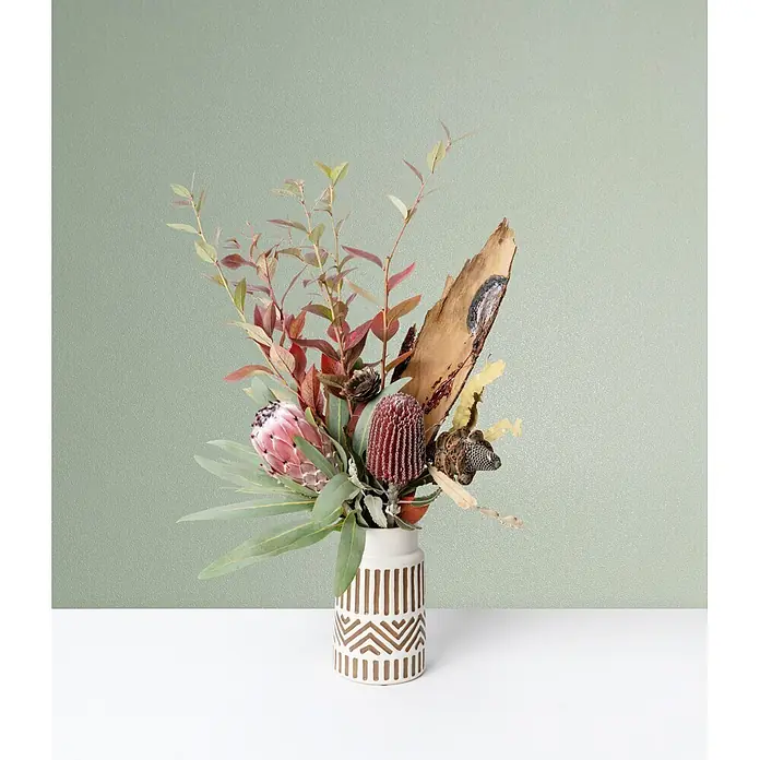 Marburg Vliestapete Novamur Naomi Uni I - Salbeigrün, Uni, 10,05 x 0,53 m - Eine weiße Vase mit braunen geometrischen Mustern enthält ein Arrangement aus Trockenblumen, darunter eine rosa Protea, rote Banksie, Blätter und ein Stück Treibholz.