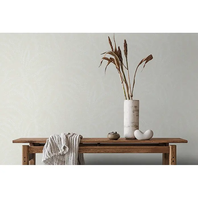 Marburg Vliestapete Novamur Oriana Floral - Weiß-Beige, Floral, 10,05 x 0,53 m - Eine Holzbank mit weißer Decke, getrockneten Pflanzen in hoher Vase und Deko-Skulpturen vor einer hell gemusterten Wand.