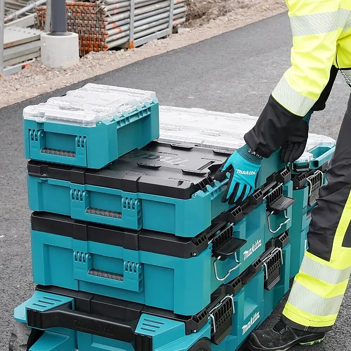 Makita Werkzeugkasten Maktrak M - Kunststoff, L x B x H: 56,2 x 38,7 x 16,2 cm - Arbeiter bewegt gestapelte, türkis-schwarze Makita Werkzeugkästen mit transparenten Deckeln auf einer Baustelle.