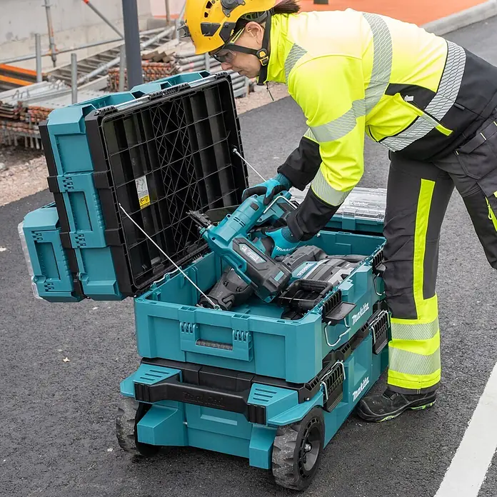 Makita Werkzeugkasten Maktrak L - Kunststoff, L x B x H: 80,2 x 56,5 x 25,2 cm - Ein Arbeiter legt Werkzeuge in ein offenes, türkis-schwarzes Makita Rollkoffersystem auf einer Baustelle.