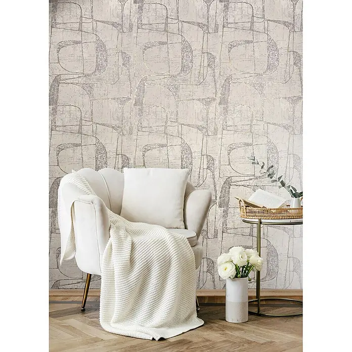 Marburg Vliestapete Novamur Leyla Grafik - Beige, Grafisch, 10,05 x 0,53 m - Abstrakte Tapete in Grau und Goldmetallic mit organischem Muster, an einer Wand in einem Wohnzimmer hinter einem cremefarbenen Sessel.
