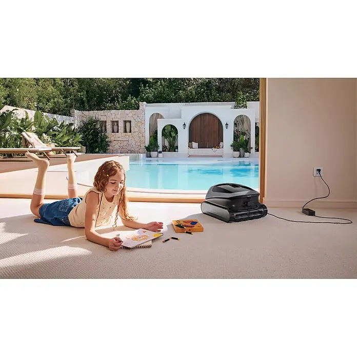 Beatbot Poolroboter Sora P3 - Akkubetrieben, Pools bis 300 m² Grundfläche, 25.700 l/h - Dunkelgrauer Poolreinigungsroboter lädt auf Teppichboden im Innenbereich mit Blick auf einen Pool.